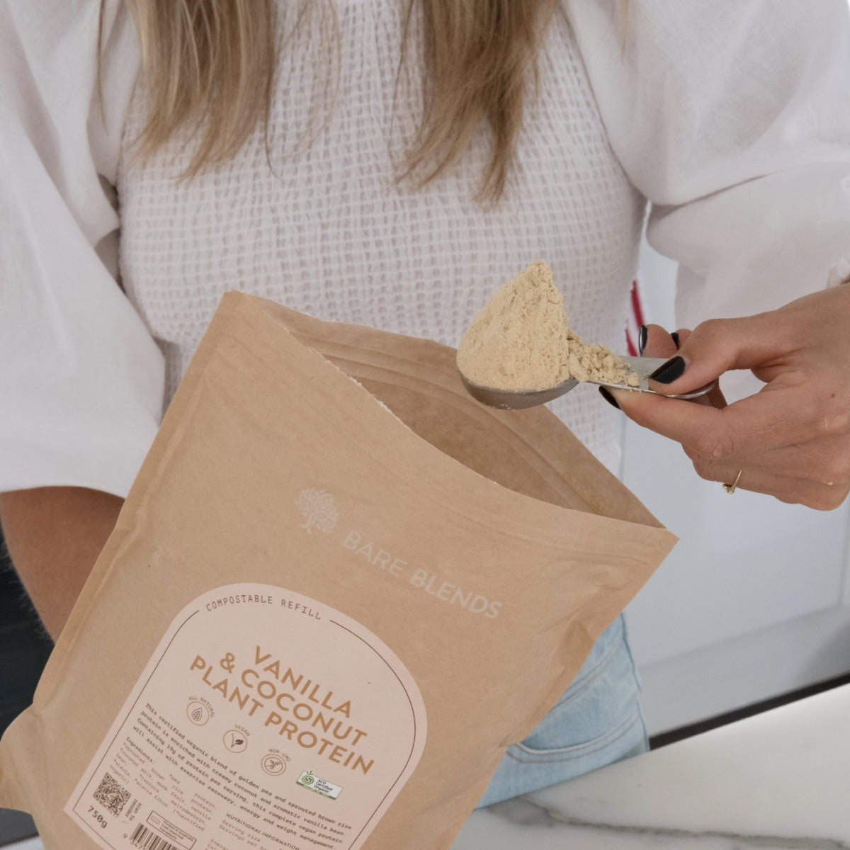 Scoop of Vanilla and Coconut Plant Protein powder held above an open Bare Blends pouch